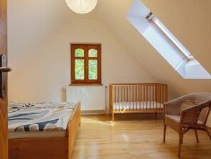 Ein Zimmer mit schrägen Wänden, einem hölzernen Wiege, einem Korbsessel, einem Bett mit gemusterten Betttextilien und einem Fenster mit Blick auf Grünflächen.