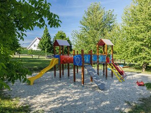 Ein Spielplatz mit Rutschen und Klettergerüsten, umgeben von Bäumen und einem Gebäude im Hintergrund.