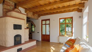 Ein geräumiges Wohnzimmer mit Kamin, Holzbalkendecke, einem großen Fenster mit Blick ins Grüne und einem weißen Sofa mit bunten Kissen.