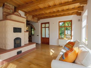 A spacious living room with a fireplace, wooden ceiling beams, a large window with a view of greenery, and a white sofa with colorful cushions. - Villa Canela by Interhome (Pécsely)