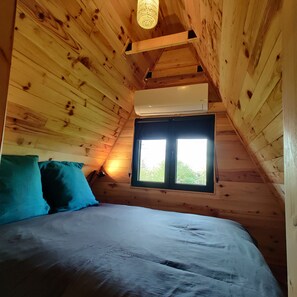 A wooden room with a bed, pillows, a window, and an air conditioning unit. - Enjoy the sounds of nature when you stay in this unique wooden Tipi. (Vivy)
