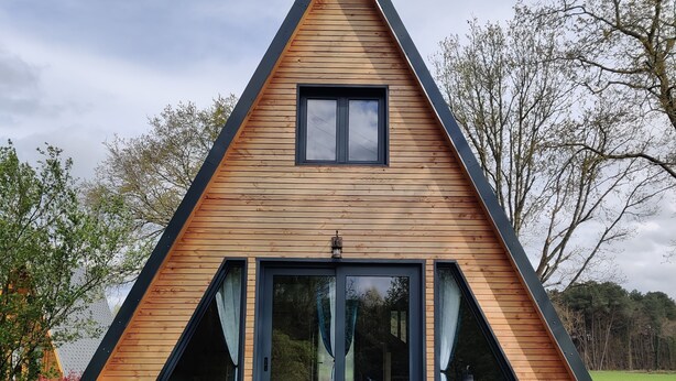 A modern A-frame wooden house with large windows, a patio with outdoor seating, and a view of the surrounding landscape.