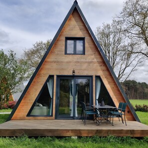 A modern A-frame wooden house with large windows, a patio with outdoor seating, and a view of the surrounding landscape. - Enjoy the sounds of nature when you stay in this unique wooden Tipi. (Vivy)