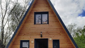 A wooden A-frame cabin with a central door, two windows on each side, and a visible air conditioning unit.