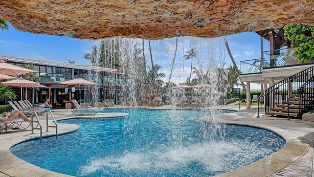 Una piscina circular con cascada, rodeada de sillas de descanso y sombrillas.