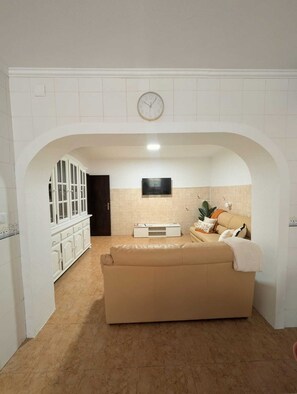 Een kamer met een beige bank, een aan de muur bevestigde televisie en een klok boven de bank.