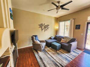 A living room with a sectional sofa, a recliner, a coffee table, a patterned rug, a ceiling fan, and a wall-mounted decorative piece.