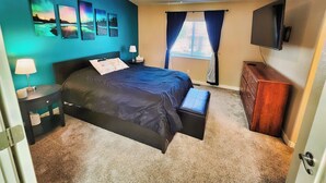 Une chambre à coucher avec un lit, un ventilateur de plafond, une télévision, une fenêtre avec des rideaux et des œuvres d’art accrochées au mur.