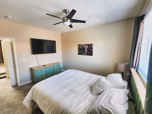 A bedroom with a bed, a ceiling fan, a TV, a dresser, and a window with curtains.