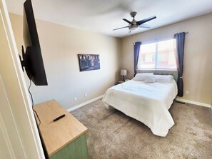 A bedroom with a bed, a ceiling fan, a painting, a lamp, a window with curtains, and a TV mounted on the wall.