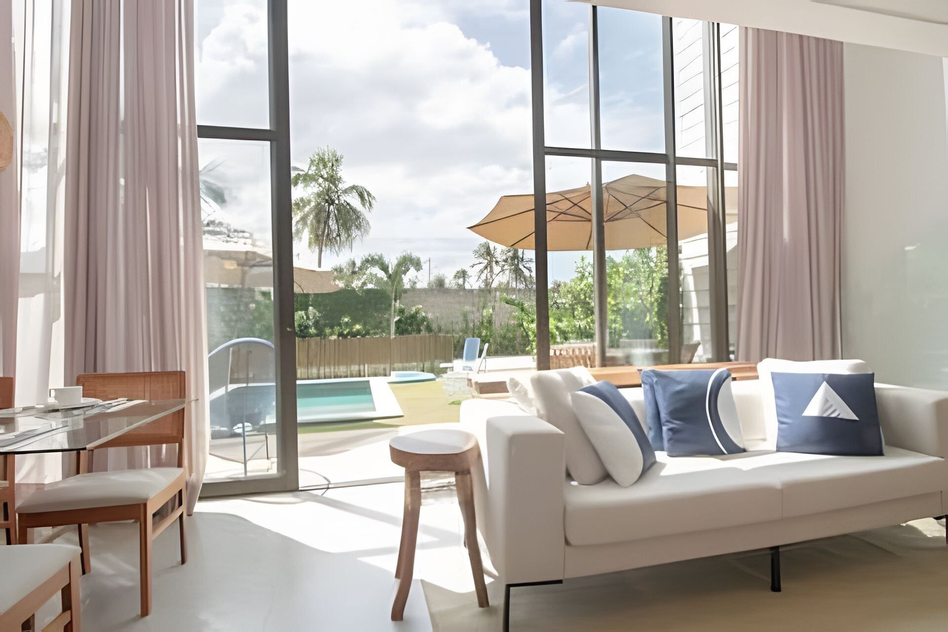 Uma sala de estar moderna com sofá, cadeiras, mesa de vidro e vista para uma piscina e palmeiras.