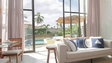 Uma sala de estar moderna com sofá, cadeiras, mesa de vidro e vista para uma piscina e palmeiras.