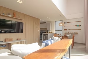 Un salon moderne avec une longue table en bois, un téléviseur fixé au mur et une cuisine en arrière-plan.