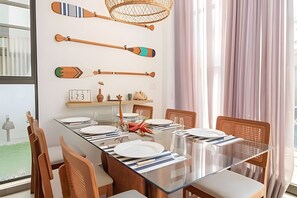 Uma sala de jantar com uma mesa comprida posta para seis pessoas, cadeiras de madeira, uma mesa com tampo de vidro e objetos decorativos na parede.
