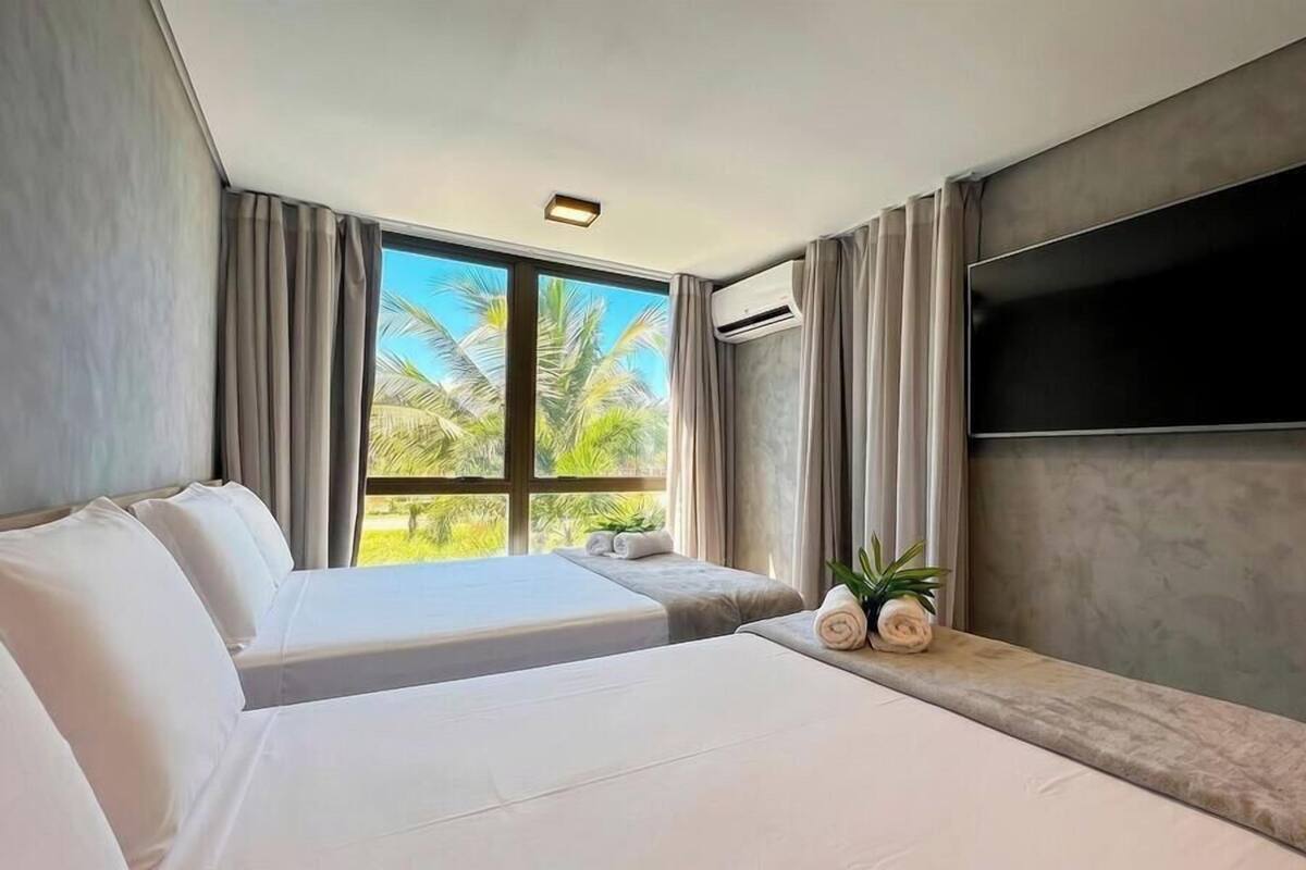 Quarto de hotel com duas camas, televisão de tela plana, aparelho de ar condicionado e janela com vista para palmeiras.