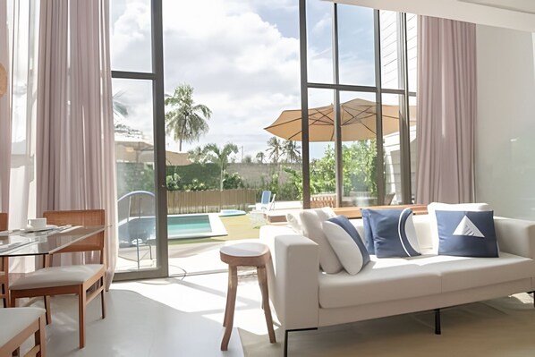 Un salon moderne avec un canapé, des fauteuils, une table en verre et une vue sur une piscine et des palmiers.