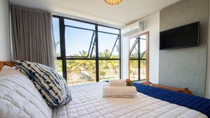 A hotel room with a large bed, a flat-screen TV, and a view of palm trees through large windows. - Oka Milagres Cond. Beira Mar By MME (Passo de Camaragibe)