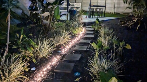 Caminho no jardim que leva a uma área coberta com mesas e cadeiras, plantas e iluminação.