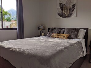 Ein Schlafzimmer mit einem Bett, einem Nachttisch, einer Lampe, einem Fenster mit Vorhängen und einem an der Wand hängenden Kunstwerk.