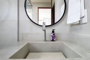 Ein Badezimmer mit einem rechteckigen Waschbecken, einem runden Spiegel und einer Handdesinfektionsflasche.