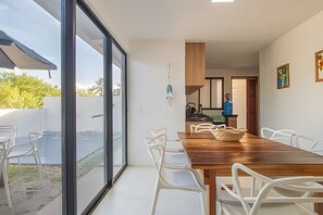 Ein moderner Essbereich mit Holztisch und weißen Stühlen, eine Schiebetür zu einem Außenbereich und eine Küche im Hintergrund.