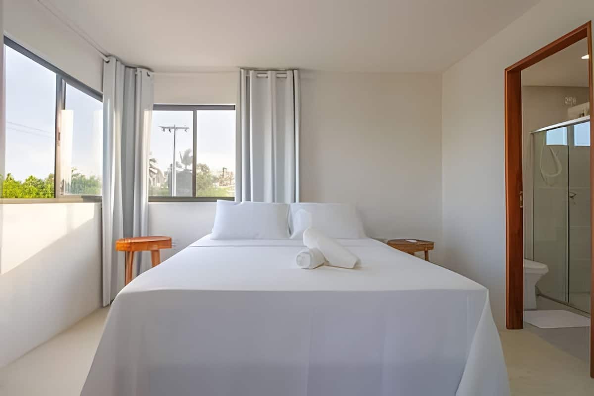 Ein Hotelzimmer mit einem großen Bett, einem kleinen Nachttisch, einem Fenster mit Vorhängen und Blick ins Freie.