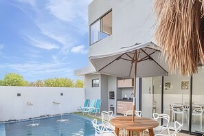 Ein moderner Poolbereich im Freien mit Esstisch und Stühlen, einer überdachten Lounge und einer Küchenzeile mit Glasfronten.