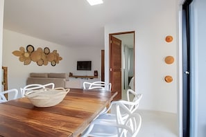 Ein Essbereich mit Holztisch, weißen Stühlen, einer gewebten Schüssel, einer Couch, einem Fernseher und einer Holztür.