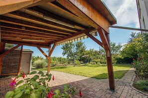 Un patio couvert avec un chemin en briques, des poutres en bois et un jardin agrémenté de pelouse et d’arbres.