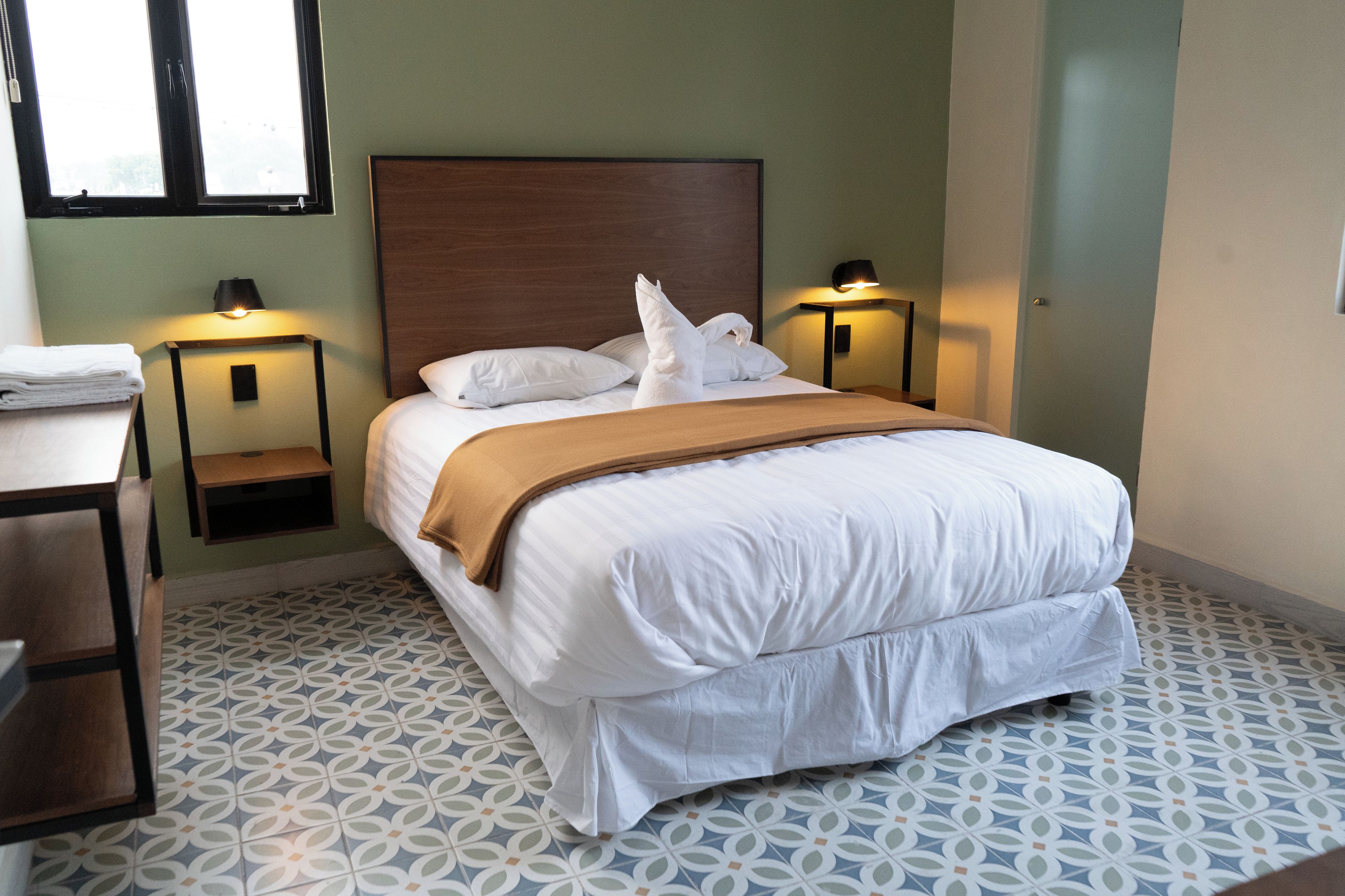 A neatly made bed with white linens and a brown throw blanket, flanked by two bedside tables with lamps, in a room with patterned floor tiles and a plain wall.