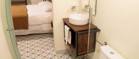 Un baño con inodoro, lavabo y espejo. El piso presenta un diseño y hay una cama en la habitación contigua.