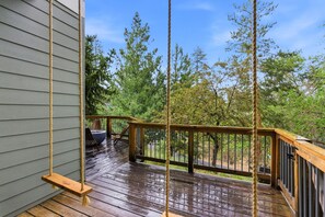 A wooden deck with a swing, a railing, and a view of trees.