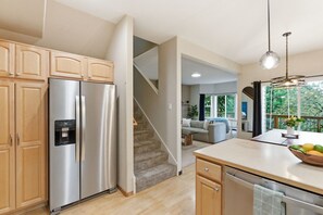 A modern kitchen with a stainless steel refrigerator, wooden cabinets, a built-in water dispenser, and a view of a living area through an open door.