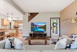 A modern living room with a large flat-screen TV, wooden furniture, and a view of a mountain lake through a sliding glass door.