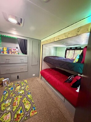 Een stapelbed met een rood en blauw matras, een klein bureau met lades, een vloerbedekking en een raam met gordijnen.