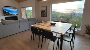 Um espaço de estar moderno com uma mesa de jantar de madeira, cadeiras pretas, um sofá cinzento, uma televisão de tela plana e uma janela ampla com vista panorâmica.