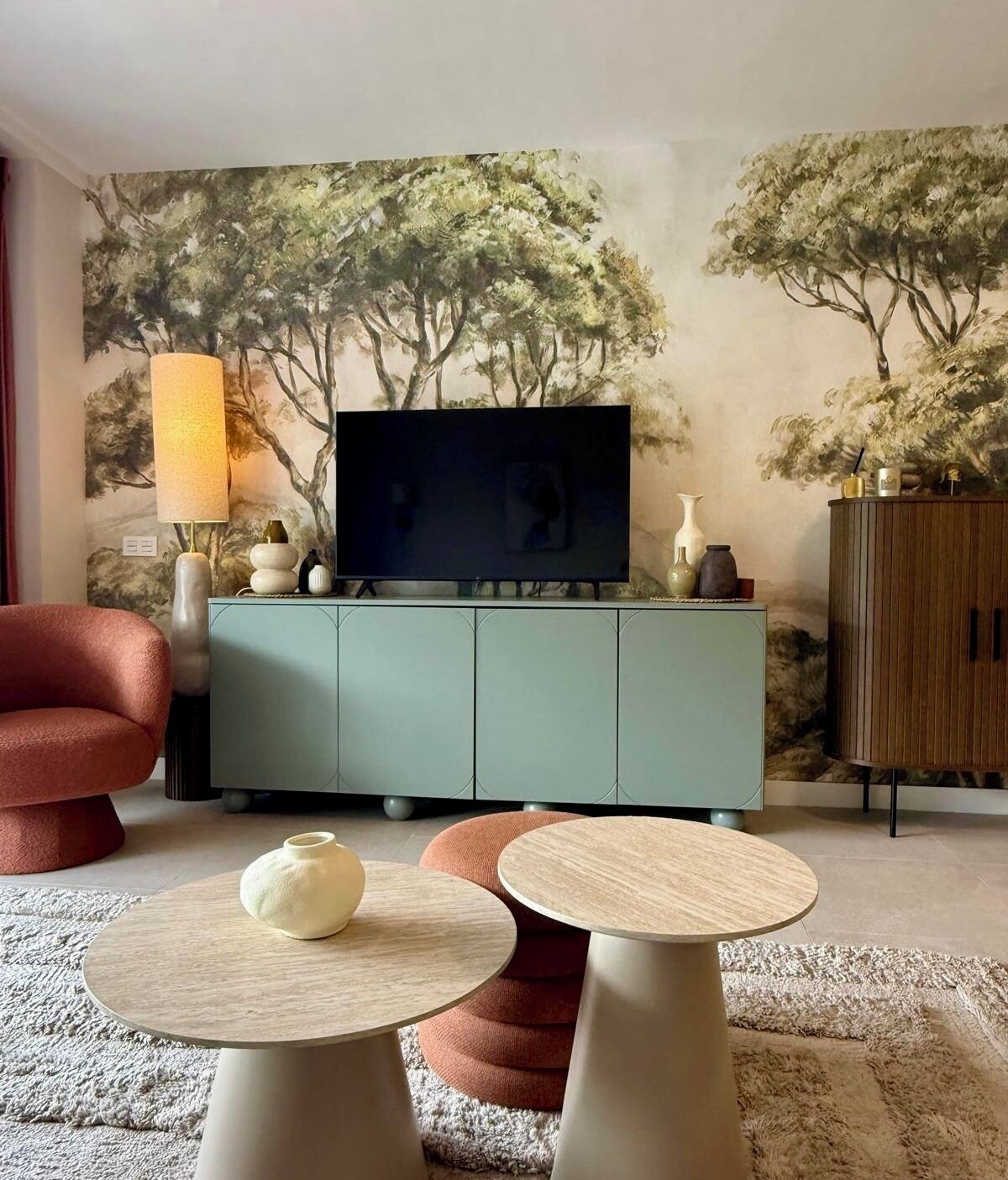 A living room with a flat-screen TV, teal cabinet, red armchair, and round wooden tables.