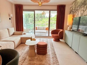 A modern living room with a sofa, a round coffee table, a red armchair, a television, and a view of a balcony with outdoor furniture.