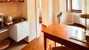 Cozinha compacta com geladeira, cafeteira e mesa de madeira com cadeiras. Há uma janela com cortinas e uma porta que dá para outro quarto.