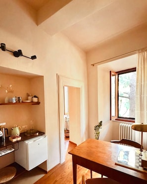Una cocina compacta con refrigerador, cafetera y una mesa de madera con sillas. Se ve una ventana con cortinas y una puerta que da a otra habitación.