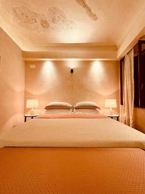 Una habitación de hotel con una cama grande, dos mesitas de noche con lámparas, un techo decorado con obras de arte y una ventana con cortinas.