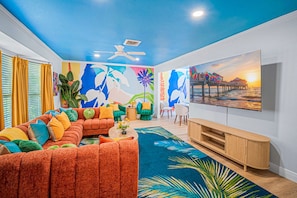 A living room with a sectional sofa, a flat-screen TV, and a vibrant mural on the wall. - Neon Tropics - Indoor Mini Golf, Pickleball, Pool (Seminole)