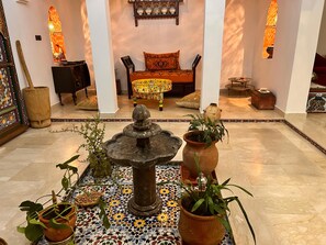 A room with a fountain, potted plants, a bed with colorful bedding, and a decorative wall. - Riad Dar Ania (fès)