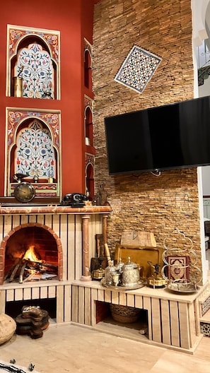 A traditional fireplace with a stone wall, a flat-screen TV mounted above it, and decorative tiles. - Riad Dar Ania (fès)