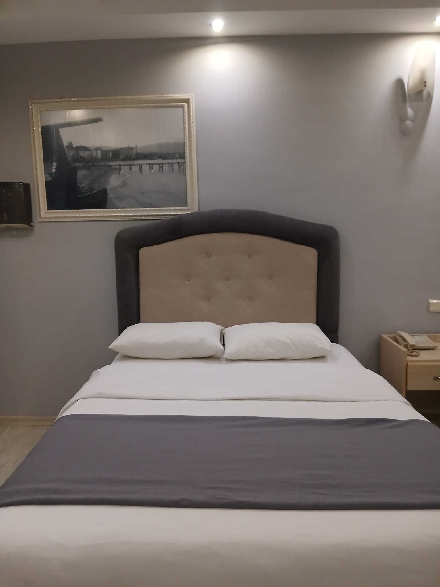 Un lit bien fait, avec une tête de lit, une table de chevet sur laquelle se trouve un téléphone et un tableau encadré accroché au mur.