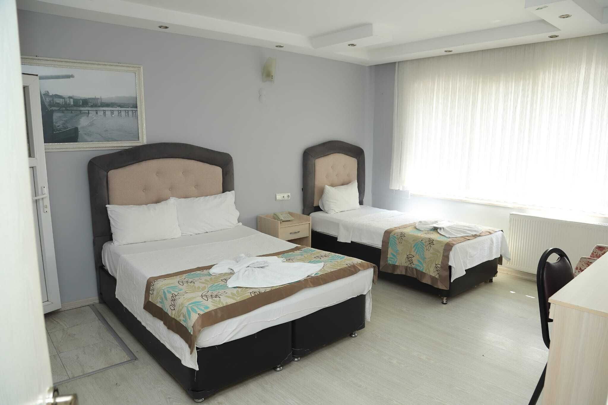 A hotel room with two beds, a desk, a chair, a window with curtains, a radiator, and a framed picture on the wall.