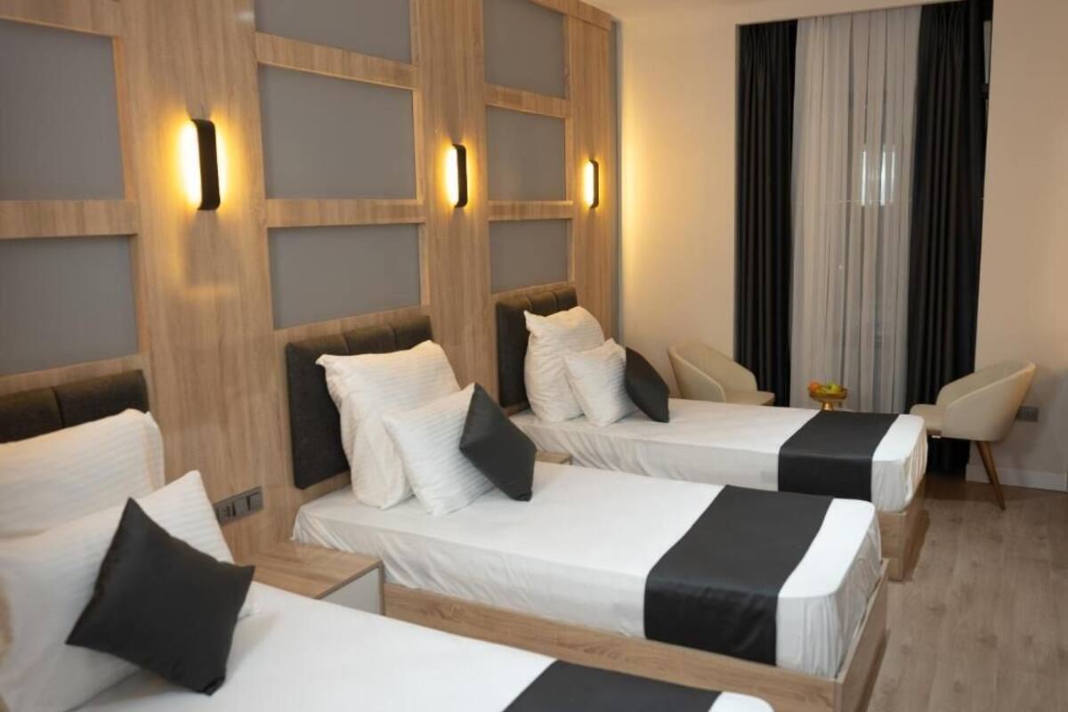 Une chambre d’hôtel avec trois lits simples, une tête de lit en bois, une petite table sur laquelle se trouve un panier de fruits et une chaise.