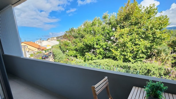 A balcony with a wooden chair and table, a potted plant, and a view of greenery and buildings.