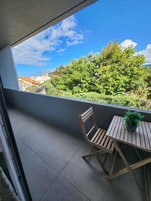 Un balcon avec une chaise et une table en bois, une plante en pot et une vue sur la verdure et les bâtiments.