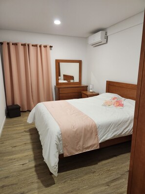 A hotel room with a bed, a wooden nightstand, a mirror, a small table, a suitcase, and an air conditioning unit. - Spacious 3-bedroom apartment with AC in welcoming Sao Miguel (Rabo de Peixe, Sao Miguel)